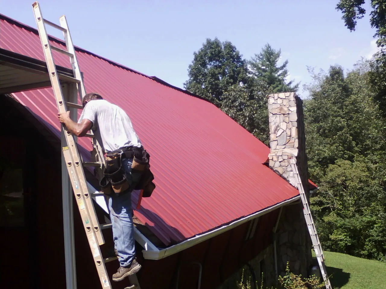 Roofer on a ladder beside a red metal roof and stone chimney — Soffit & Fascia Repair by Roofing Brothers in The Dalles